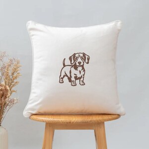 May include: A square, off-white decorative pillow featuring a brown illustration of a dachshund dog. The dog is standing, facing forward, with its tail curled up. The pillow is placed on a wooden stool.