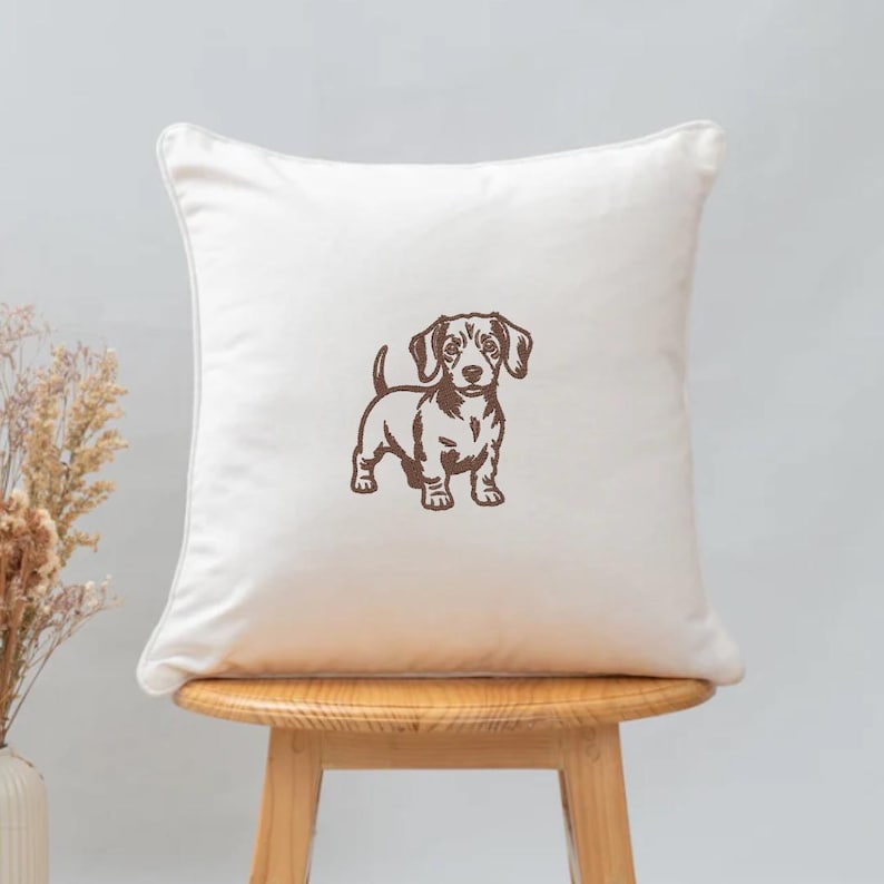 May include: A square, off-white decorative pillow featuring a brown illustration of a dachshund dog. The dog is standing, facing forward, with its tail curled up. The pillow is placed on a wooden stool.