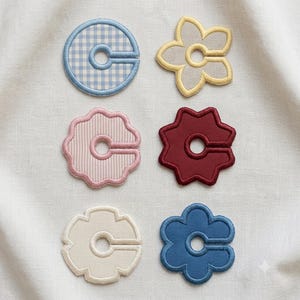 Peut inclure: Six écussons brodés en forme de lettre C, chacun avec une découpe circulaire centrale. Les motifs incluent un cercle à carreaux bleus, une fleur jaune, une forme festonnée à rayures roses, une étoile rouge foncé, une fleur crème et une fleur bleue.