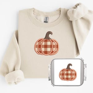 Peut inclure: Un sweat-shirt à col rond beige avec un motif de citrouille brodé au centre, présentant un motif à carreaux vichy marron et blanc. La citrouille a une tige marron foncé. Un cadre à broder assorti est placé dans le coin inférieur droit.
