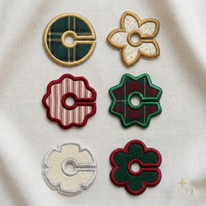 Peut inclure: Six séparateurs de vêtements en tissu brodé avec une découpe circulaire centrale et une fente. L'ensemble comprend un cercle à carreaux vert, une fleur crème à motifs, une forme festonnée à rayures rouges, une étoile à carreaux vert et rouge.