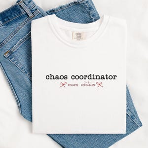 May include: A white t-shirt with the words "chaos coordinator mom edition" printed in black and burgundy. The shirt is folded and placed on top of a pair of blue jeans.