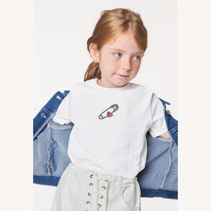 Peut inclure: Un t-shirt blanc avec un motif d'épingle à nourrice et de cœur rouge. Le t-shirt est porté avec une veste en jean et un short blanc. L'image est prise en studio.