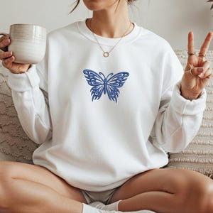 May include: White crew neck sweatshirt with a detailed blue butterfly embroidered on the front. The butterfly design features intricate patterns and a symmetrical wing structure. The sweatshirt has long sleeves and a relaxed fit, suitable for casual wear.