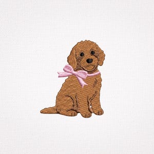 Peut inclure: Un écusson brodé représentant un chiot brun doré assis. Le chiot a des yeux brun foncé, un nez noir et un ruban rose clair noué autour du cou. La broderie utilise du fil texturé pour créer l'apparence d'un pelage poilu.