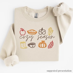 Peut inclure: Un sweat-shirt beige à col rond avec des broderies sur le thème de l'automne, notamment une pomme, une tarte, des glands, du maïs, des bottes de pluie, une tasse, une citrouille et une feuille de chêne. Le texte "cozy season" est brodé en cursif.