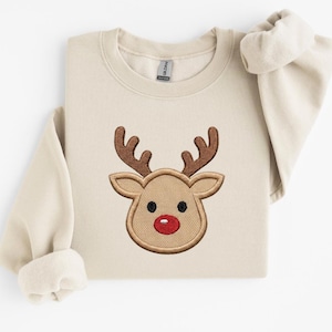 Peut inclure: Un sweat-shirt à col rond beige avec un motif brodé centré représentant une tête de renne. Le renne a un pelage brun clair, des bois brun foncé, des yeux noirs circulaires et un nez rouge vif. Le vêtement a des manches longues et un col côtelé.