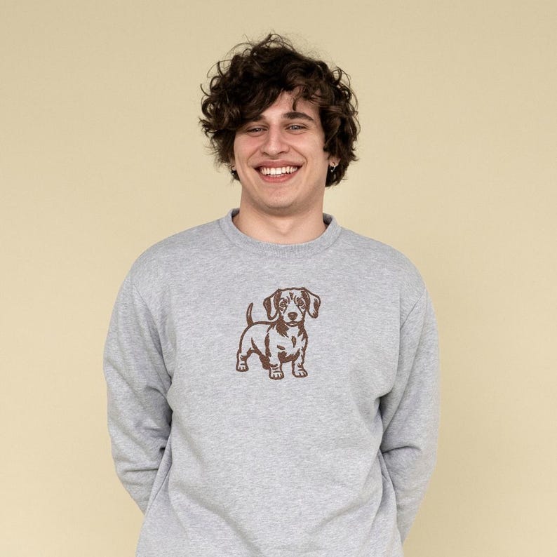 May include: A gray sweatshirt featuring a brown dachshund graphic. The long-sleeved crew neck top is made of a soft material. The dog graphic is a detailed illustration of a dachshund.