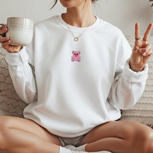 May include: White sweatshirt with a small pink teddy bear graphic on the chest. The bear has a smiling face and is embroidered with a textured design. The sweatshirt has a crew neck and long sleeves.