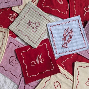 Peut inclure: Une collection de dessous de verre carrés en tissu avec des motifs brodés incluant un homard rouge, des cerises, un morceau de fromage et la lettre M. Chaque dessous de verre présente une bordure festonnée cousue d'un fil contrastant.