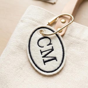 May include: A cream-coloured tote bag with a leather handle and a gold-coloured clasp. An oval, off-white tag with the letters "CM" embroidered in black is attached to the bag. The bag is made of canvas.