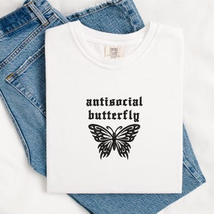 May include: White t-shirt with the text "antisocial butterfly" in a gothic font above a black butterfly graphic. The shirt is folded on top of blue denim jeans.