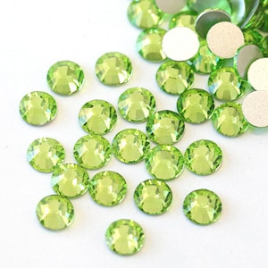 May include: A large assortment of lime green rhinestones, also known as glass gems, are scattered on a white surface. The rhinestones are round and have a faceted design.