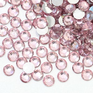 May include: A scattering of light pink, round rhinestones with a flat, silver backing. The rhinestones are spread across a white surface, with varying sizes and a sparkling appearance. Suitable for crafts and embellishments.