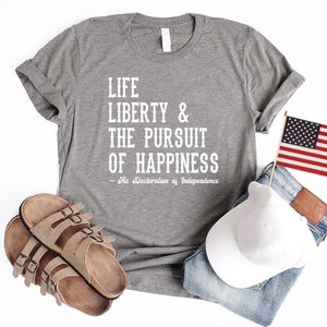 May include: A heather grey t-shirt with the text "Life Liberty & The Pursuit of Happiness - The Declaration of Independence" printed in white. The shirt is laid flat on a white surface with a pair of brown sandals, a white baseball cap, and a small American flag.
