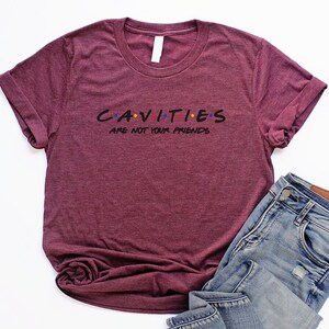 Może przedstawiać: Bordowy t-shirt z napisem "CAVITIES ARE NOT YOUR FRIENDS" w czarnym kolorze z kolorowymi kropkami. Tekst jest stylizowany na logo "Friends".