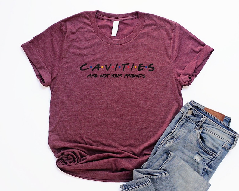 Może przedstawiać: Bordowy t-shirt z napisem "CAVITIES ARE NOT YOUR FRIENDS" w czarnym kolorze z kolorowymi kropkami. Tekst jest stylizowany na logo "Friends".