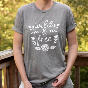 Op de afbeelding: Een heidegrijs T-shirt met een witte afbeelding waarop "wild & free" staat met bloemmotieven.