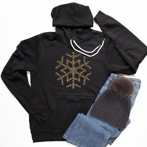 May include: Black hooded sweatshirt with a gold snowflake design featuring a leopard print pattern. The sweatshirt is paired with blue jeans and a dark gray beanie with a fluffy pom-pom.