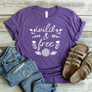 Op de afbeelding: Paars T-shirt met de tekst "wild & free" in witte letters met bloemmotieven. Het shirt ligt op een houten oppervlak met een paar bruine sandalen.