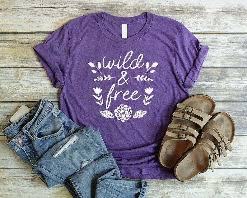 Op de afbeelding: Paars T-shirt met de tekst "wild & free" in witte letters met bloemmotieven. Het shirt ligt op een houten oppervlak met een paar bruine sandalen.