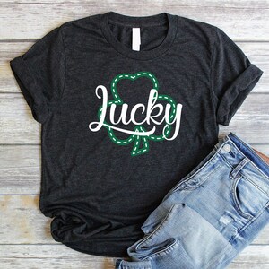 May include: A dark gray t-shirt with a white graphic of a shamrock with the word "lucky" inside. The shirt is folded and laid on a wooden surface with a pair of blue jeans.