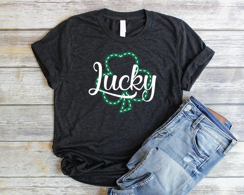 May include: A dark gray t-shirt with a white graphic of a shamrock with the word "lucky" inside. The shirt is folded and laid on a wooden surface with a pair of blue jeans.