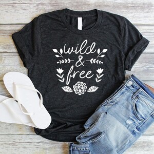 Op de afbeelding: Een donkergrijs T-shirt met de tekst "wild & free" in witte letters. De tekst wordt omringd door kleine witte bloemenmotieven. Het shirt ligt plat op een houten oppervlak met een paar witte slippers en een blauwe spijkerbroek.