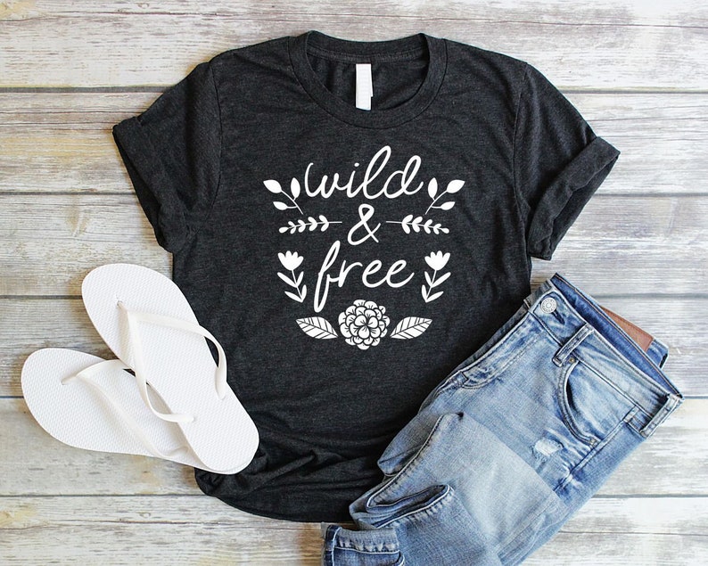 Op de afbeelding: Een donkergrijs T-shirt met de tekst "wild & free" in witte letters. De tekst wordt omringd door kleine witte bloemenmotieven. Het shirt ligt plat op een houten oppervlak met een paar witte slippers en een blauwe spijkerbroek.