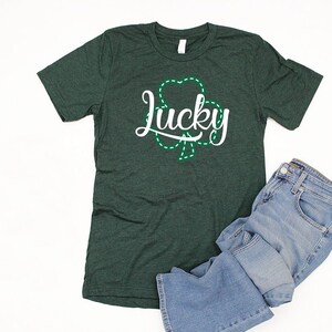 May include: A dark green t-shirt with a white graphic of a shamrock and the word "lucky" in a white cursive font.