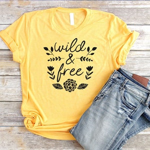 Op de afbeelding: Een geel T-shirt met zwarte tekst die "Wild & Free" zegt met bloemmotieven. Het shirt is gevouwen en ligt op een houten oppervlak met een blauwe spijkerbroek.