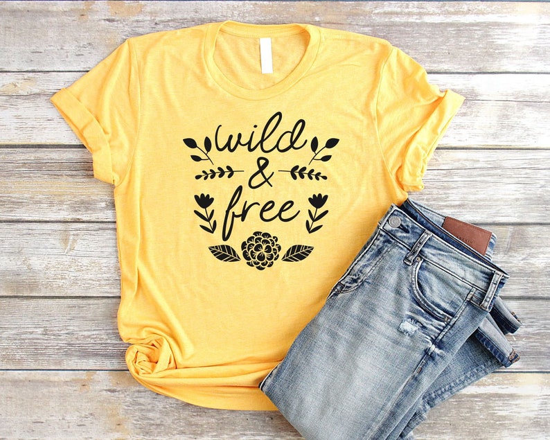 Op de afbeelding: Een geel T-shirt met zwarte tekst die "Wild & Free" zegt met bloemmotieven. Het shirt is gevouwen en ligt op een houten oppervlak met een blauwe spijkerbroek.