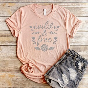 Op de afbeelding: Een perzikkleurig T-shirt met de tekst "wild & free" in grijze letters met bloemmotieven. Het shirt ligt op een houten oppervlak met een spijkerbroek.