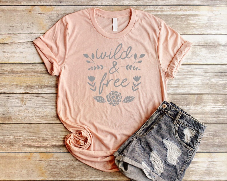 Op de afbeelding: Een perzikkleurig T-shirt met de tekst "wild & free" in grijze letters met bloemmotieven. Het shirt ligt op een houten oppervlak met een spijkerbroek.