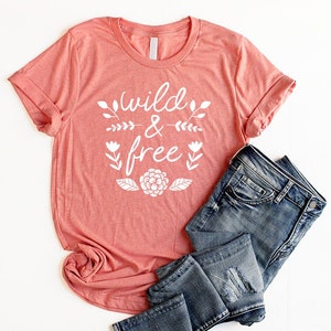 Op de afbeelding: Een koraalkleurig T-shirt met de tekst "wild & free" in witte letters met bloemmotieven. Het shirt is gevouwen en wordt getoond met een blauwe spijkerbroek.