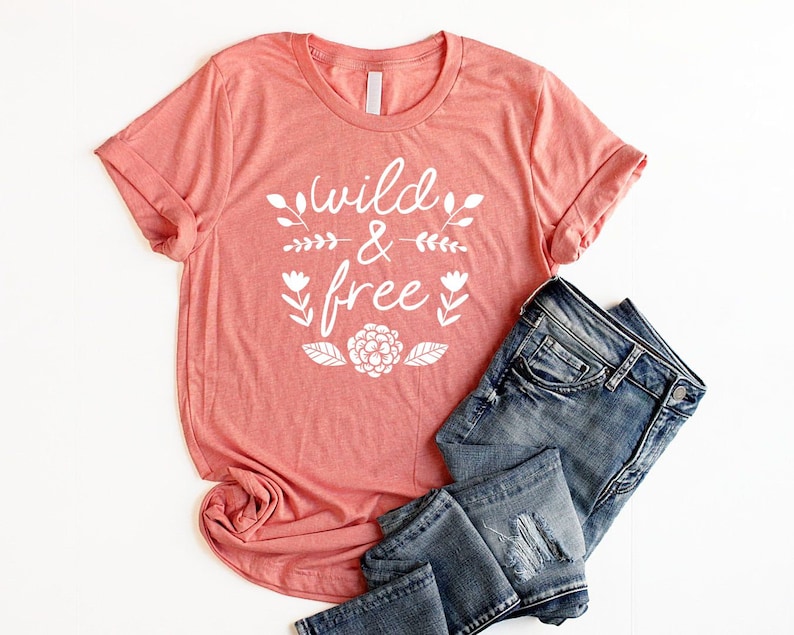 Op de afbeelding: Een koraalkleurig T-shirt met de tekst "wild & free" in witte letters met bloemmotieven. Het shirt is gevouwen en wordt getoond met een blauwe spijkerbroek.