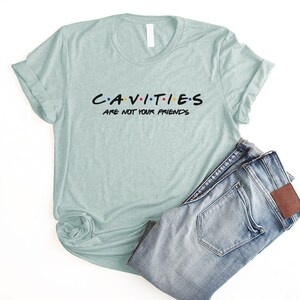 Może przedstawiać: Jasno błękitny t-shirt z napisem "CAVITIES ARE NOT YOUR FRIENDS" w kolorze czarnym z kolorowymi kropkami. Koszulka jest złożona i leży na białym tle z niebieskimi dżinsami.
