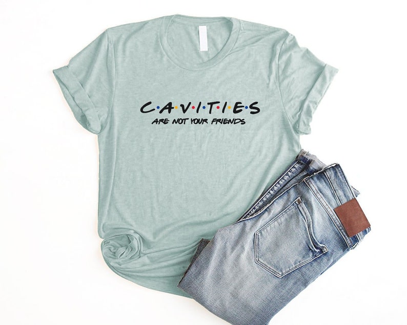 Może przedstawiać: Jasno błękitny t-shirt z napisem "CAVITIES ARE NOT YOUR FRIENDS" w kolorze czarnym z kolorowymi kropkami. Koszulka jest złożona i leży na białym tle z niebieskimi dżinsami.