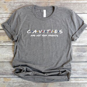 Może przedstawiać: Szary T-shirt z napisem "CAVITIES ARE NOT YOUR FRIENDS" w stylizowanym foncie z kolorowymi kropkami. Tekst jest grą słowną nawiązującą do popularnego serialu telewizyjnego "Przyjaciele".