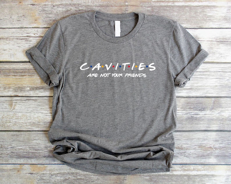 Może przedstawiać: Szary T-shirt z napisem "CAVITIES ARE NOT YOUR FRIENDS" w stylizowanym foncie z kolorowymi kropkami. Tekst jest grą słowną nawiązującą do popularnego serialu telewizyjnego "Przyjaciele".