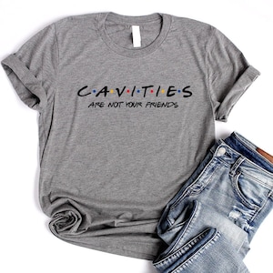 Może przedstawiać: Szara koszulka z nadrukiem "CAVITIES ARE NOT YOUR FRIENDS" w stylizowanym foncie przypominającym logo serialu "Przyjaciele". Tekst jest czarny z kolorowymi kropkami.