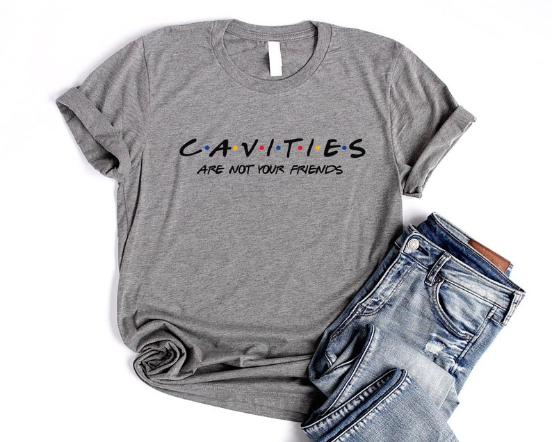 Może przedstawiać: Szara koszulka z nadrukiem "CAVITIES ARE NOT YOUR FRIENDS" w stylizowanym foncie przypominającym logo serialu "Przyjaciele". Tekst jest czarny z kolorowymi kropkami.