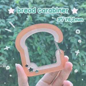 Könnte beinhalten: Ein brotförmiger Karabiner in hellorange und durchscheinendem Weiß. Der Artikel verfügt über kleine sternförmige Ausstanzungen und trägt den Text bread carabiner sowie 3" / 76,2mm. Er besitzt einen metallenen Federverschluss zum Öffnen.