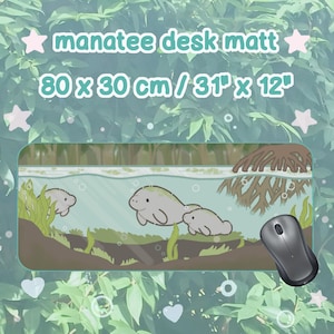 Op de afbeelding: Een rechthoekige bureaumat met een cartoonillustratie van zeekoeien die in het water zwemmen tussen waterplanten en mangrovewortels. De tekst luidt manatee desk matt 80 x 30 cm / 31 x 12. Een zwarte computermuis bevindt zich aan de rechterkant.