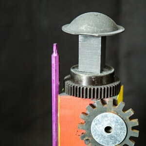 Op de afbeelding: Een paarse schroevendraaier staat naast een sculptuur gemaakt van metalen tandwielen, een grijze blok en een grijze koepel. De sculptuur is geschilderd met rode, oranje en gele verf.