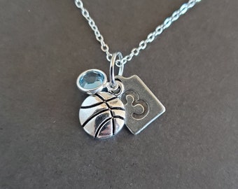 May include: Silver necklace with a basketball charm, a blue gemstone, and a tag with the number 3. The basketball charm has detailed lines. The necklace is on a dark gray background.