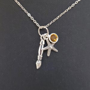 Può includere: Collana d'argento con tre ciondoli: un pennello, una stella marina e una pietra rotonda color ambra. La catena è sottile e i ciondoli sono dettagliati. Lo sfondo è nero pieno.