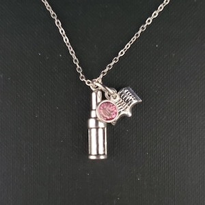 Può includere: Collana d'argento con tre ciondoli: un flacone di profumo, una gemma rosa e un piccolo libro. La collana è appesa su uno sfondo scuro. Il ciondolo del flacone di profumo è cilindrico.