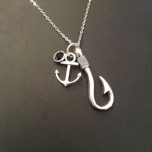 May include: Silver-toned necklace featuring a fishing hook charm, an anchor charm, and a small black gemstone. The chain is delicate, and the charms are detailed. The necklace is displayed against a dark background.