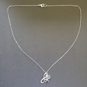 May include: A silver-colored necklace with a delicate chain and a pendant. The pendant features a stylized letter "A" intertwined with a small figure, possibly representing a person or a symbol of connection. The necklace is displayed against a dark background.
